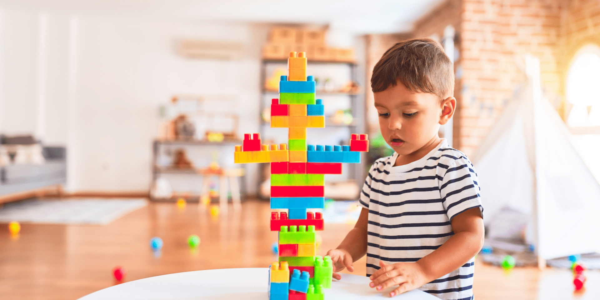 A young child in a striped shirt is focused on building a colorful structure with interlocking plastic blocks on a white table in a playroom.
