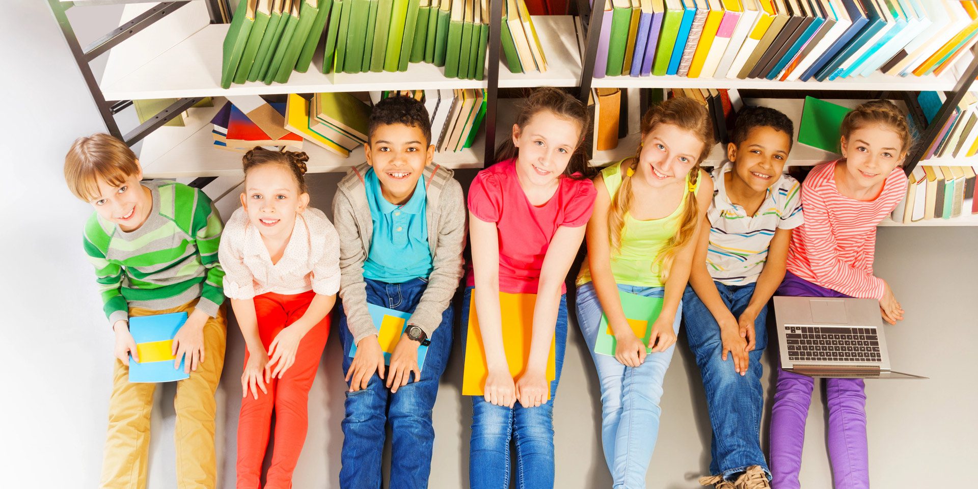 Four children sitting in a library holding books and smiling.
